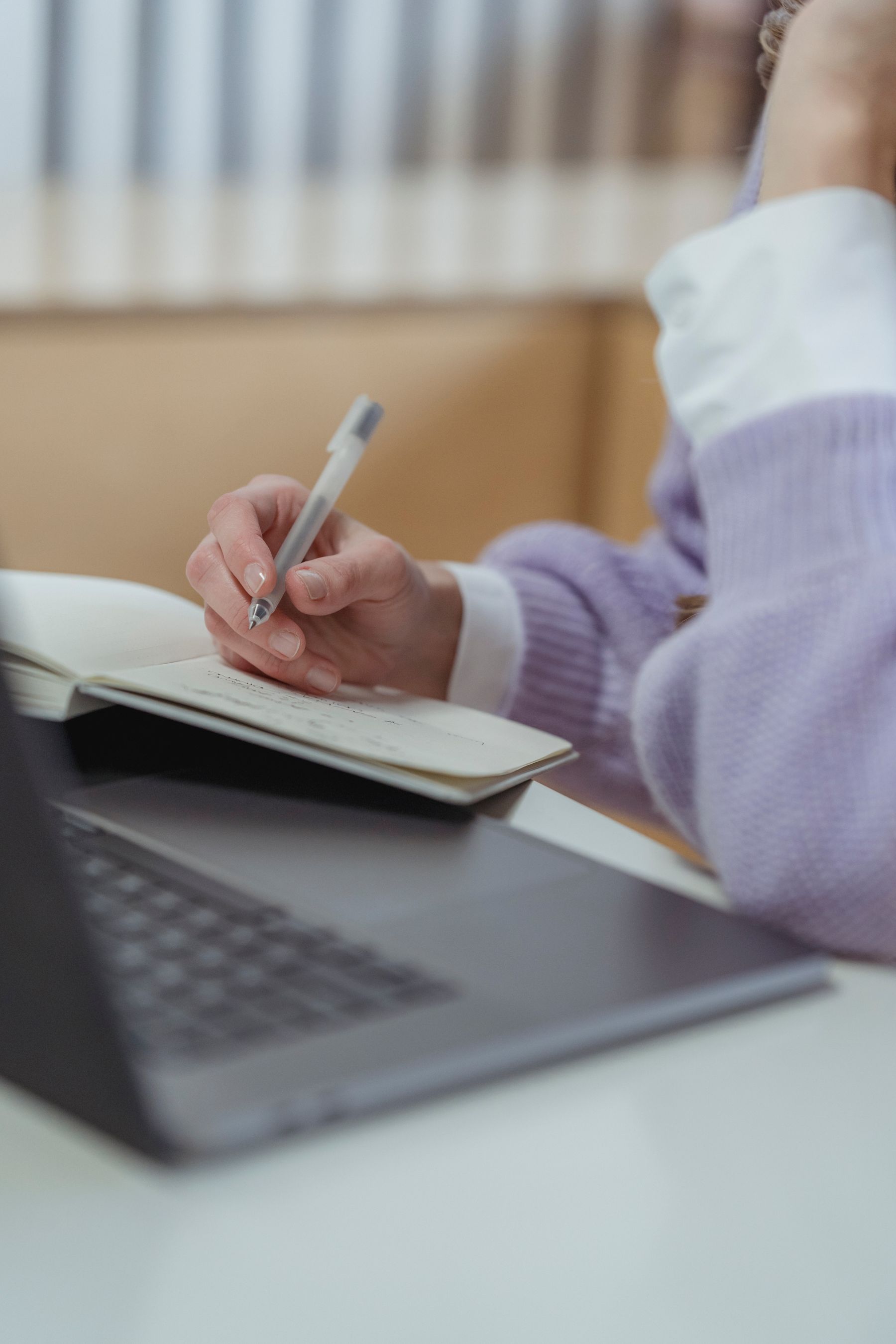 A hand writing notes in a notebook beside a laptop
